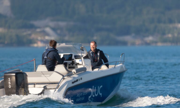 Two people are on a blue and white motorboat speeding across a lake. The boat is named 