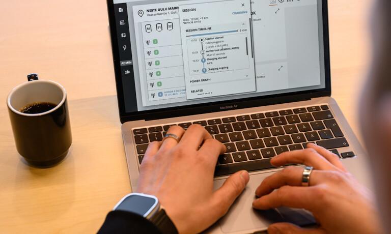 Person using a laptop with a an ev charging software program open on the screen, a cup of coffee nearby, and wearing a smartwatch on their left wrist.