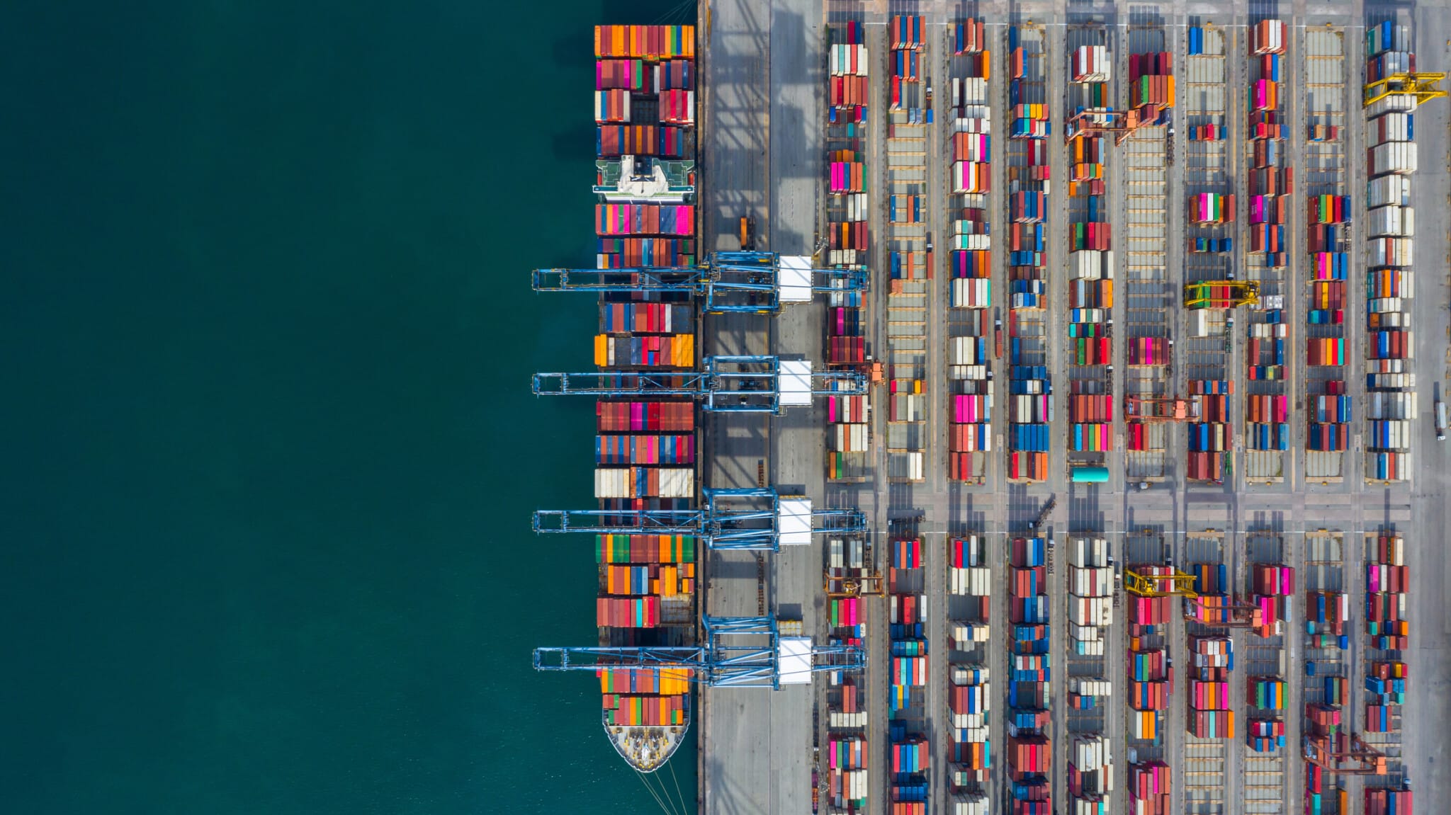 Aerial view of a bustling cargo port with multiple cranes loading and unloading colorful containers from a docked ship, depicting vibrant trade activity.