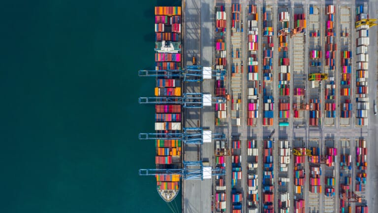 Aerial view of a bustling cargo port with multiple cranes loading and unloading colorful containers from a docked ship, depicting vibrant trade activity.