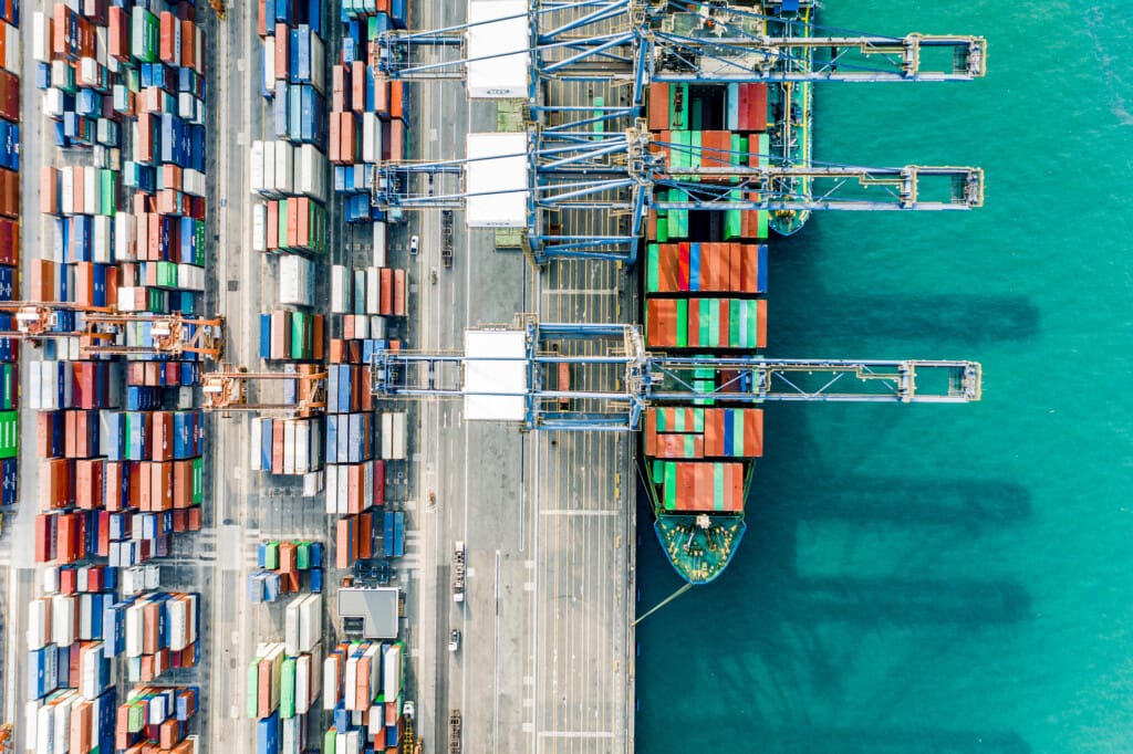 Aerial view of a bustling cargo port with multiple cranes loading and unloading colorful containers from a docked ship, depicting vibrant trade activity.