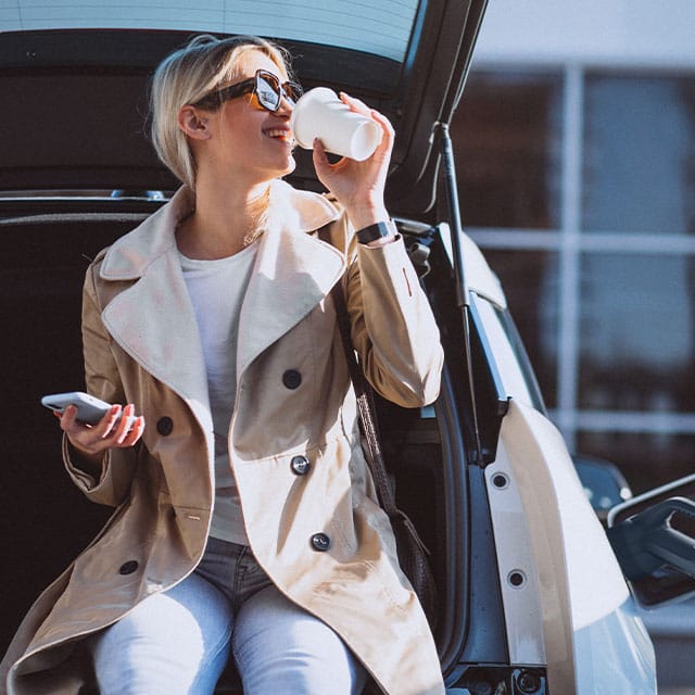 A woman in sunglasses and a beige coat sits on the trunk of a car, drinking from a paper cup and holding a smartphone in her other hand, enjoying the sunshine.