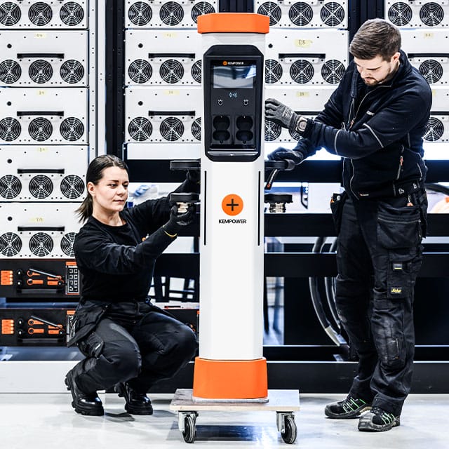 Two technicians work on installing a white and orange electric vehicle charging station indoors. The man stands on the right, using a tool on the unit, while the woman kneels on the left, adjusting another component. Server racks are visible in the background.