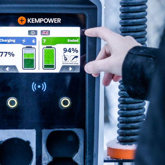 A person is pointing at a display screen on a Kempower electric vehicle charging station. The screen indicates that the charging session has ended, with the battery charged to 94%. The station has two charging ports below the screen and a coiled cable on the side.