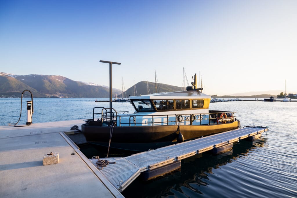 A ferry boat docked at a jetty in a calm lake with mountains in the background under a clear blue sky during sunset.