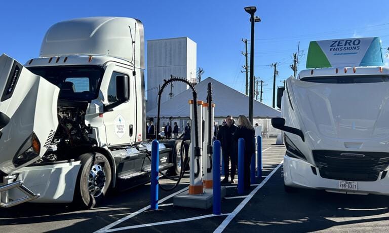 Two large white trucks with open hoods are parked side by side at a charging station, with several people standing in between under a clear blue sky.