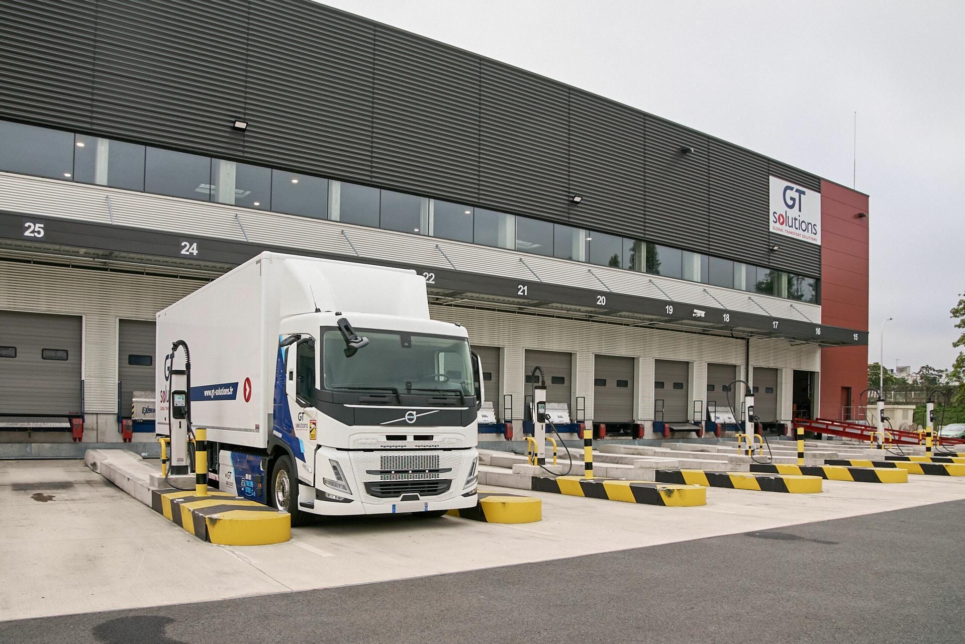 A white delivery truck is parked at a loading dock in front of a large warehouse building with multiple bay doors. The building has 